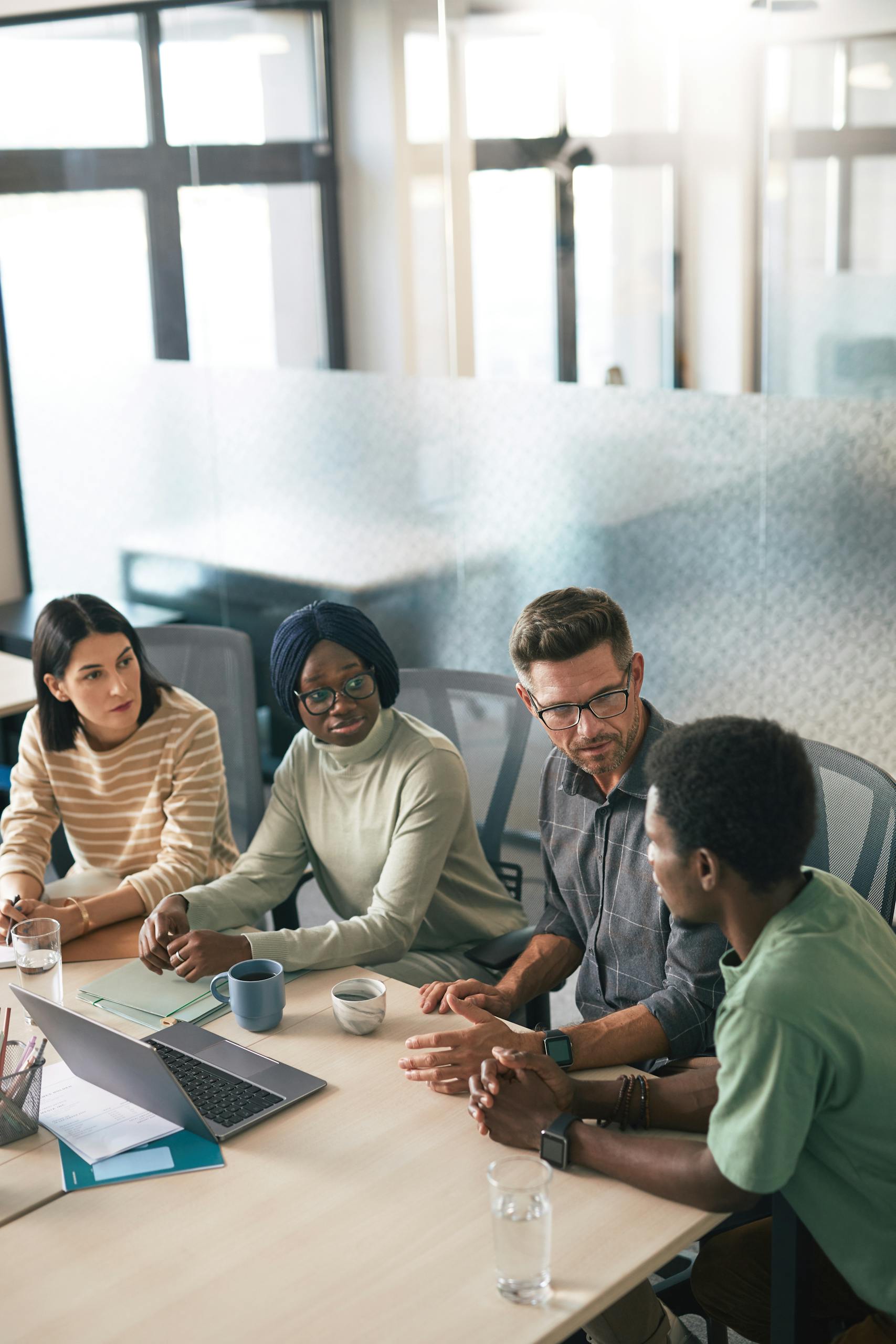 A diverse group of colleagues having a discussion in a modern office setting with laptops and coffee cups.