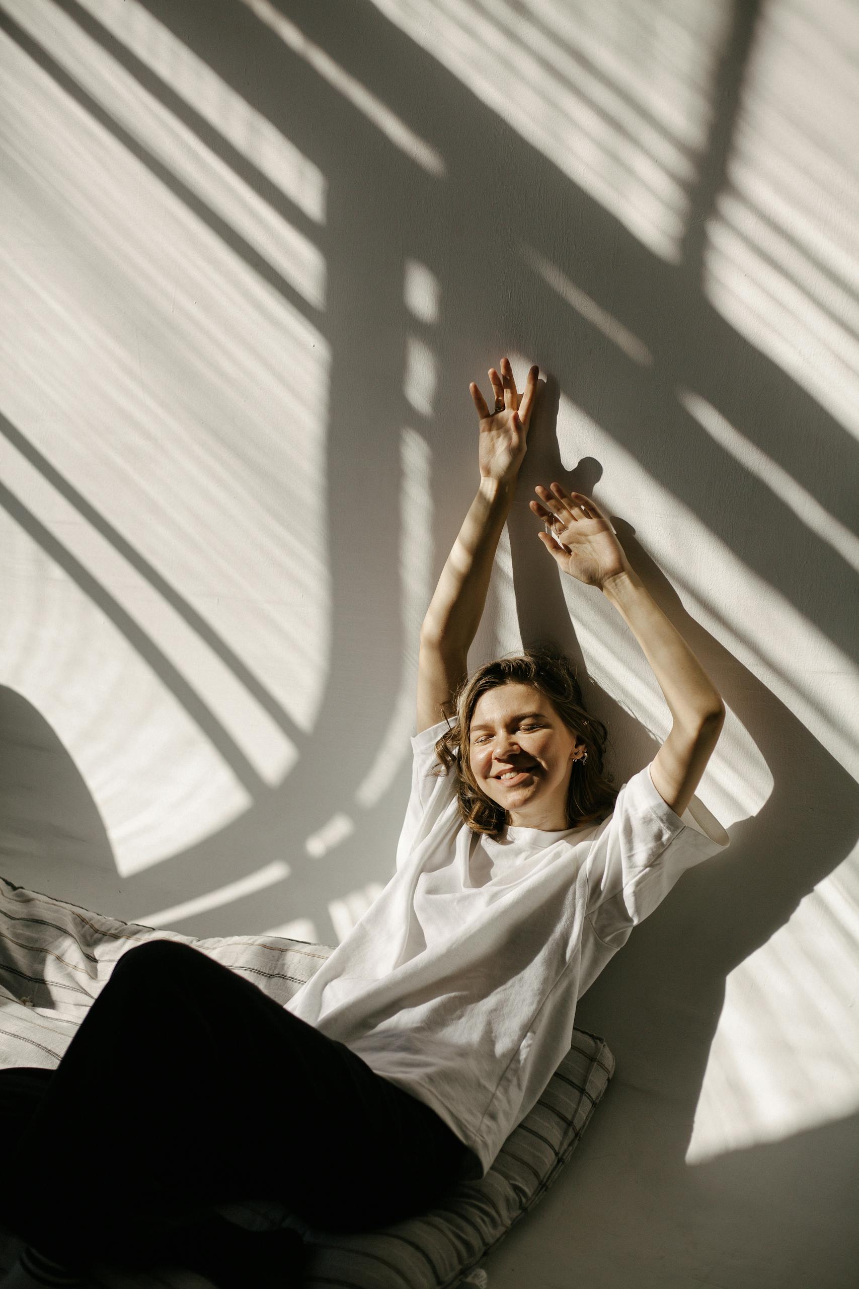 Smiling woman stretching with arms up in sunlight and shadows indoors.